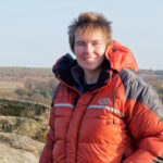 Image of woman wearing burnt orange colour down jacket with peak district rocks in background