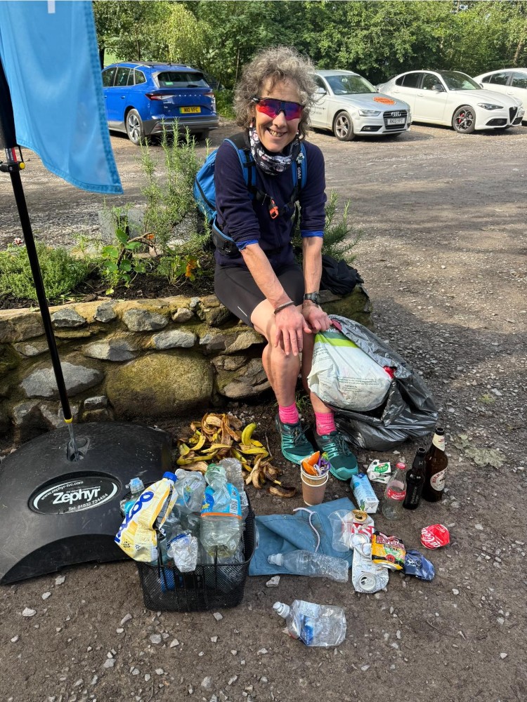 A woman in running shoes and running kit sits on a wall with a bin bag of rubbish, and other items of litter displayed at her feet