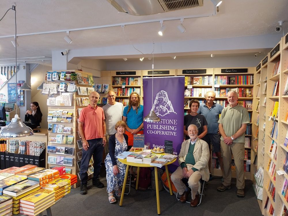 In a book shop, all eight members either standing or sitting in a semicircle in front of the Gritstone banner and behind a desk displaying books recently published by Gritstone