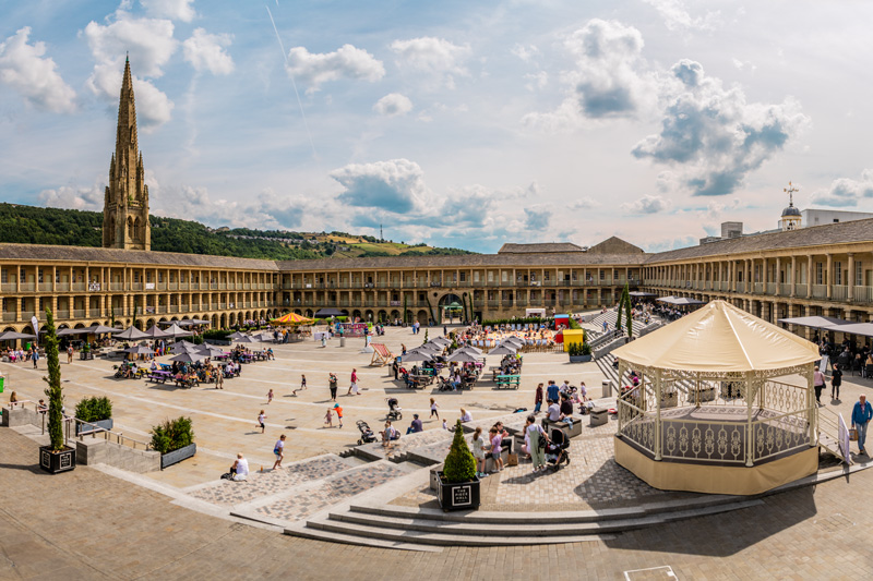 Image of Piece Hall showing bandstand, in large paved courtyard with large Gothic columned building in background, under blue skies